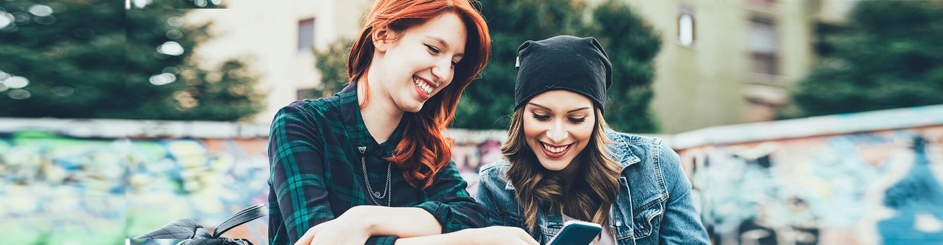 Programa Joven Up - Dos chicas jóvenes sonriendo mientras están con el móvil en la mano sentadas en un parque
