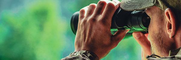 Seguros del Cazador - Cazador con prismaticos en las manos y con gorra y ropa de camuflaje en el bosque Seguros del Cazador - Cazador con prismaticos en las manos y con gorra y ropa de camuflaje en el bosque