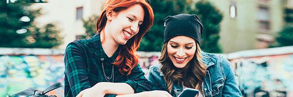 Programa Joven Up - Dos chicas jovenes sonriendo mientras navegan por intenet con el movil en un parque