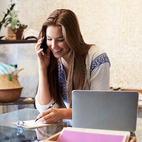 Seguros de Ahorro Particulares - Joven mujer sonriendo mientras habla por el móvil y escribe en un papel junto a un portátil sentada en el salón de su casa