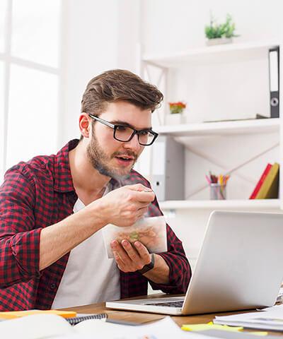 Porque somos asi Nomina - Chico joven con una camisa de cuadros rojos y gafas comiendo y trabajando frente a un portatil en la oficina